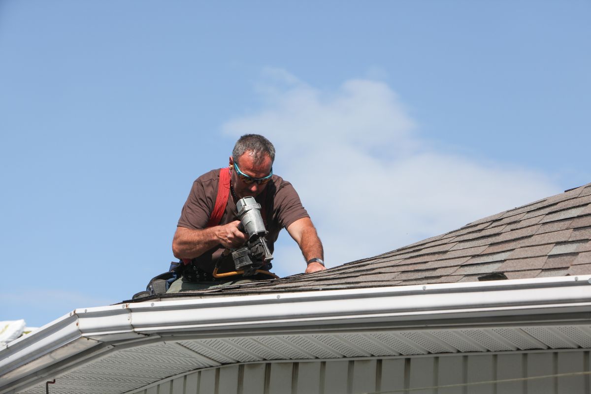 A man using a nail gun to secure roofing materials while working on a sloped roof under clear blue skies.